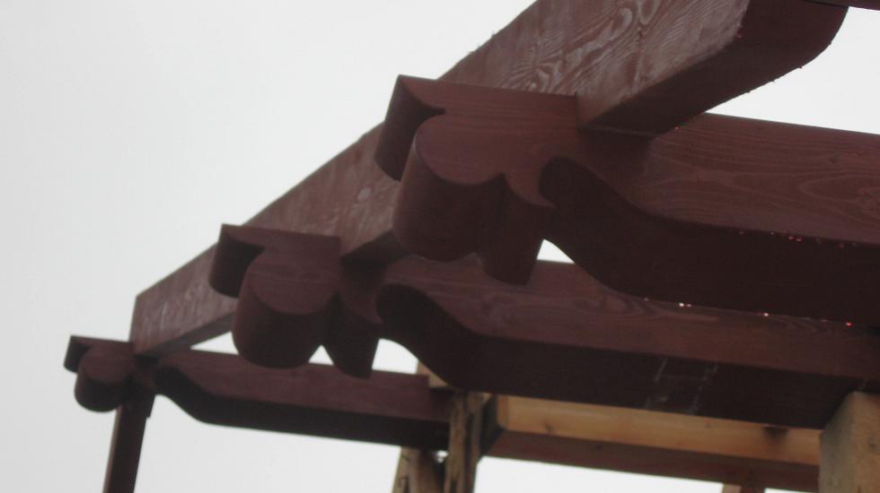 Drewniana konstrukcja pergoli z ozdobnymi wycięciami, pomalowana na ciemnobrązowy kolor, widok od dołu w pochmurny dzień.